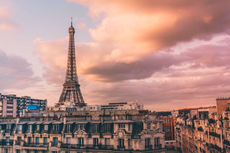 The Eiffel Tower standing above Parisian roofs against a pink and orange sky.