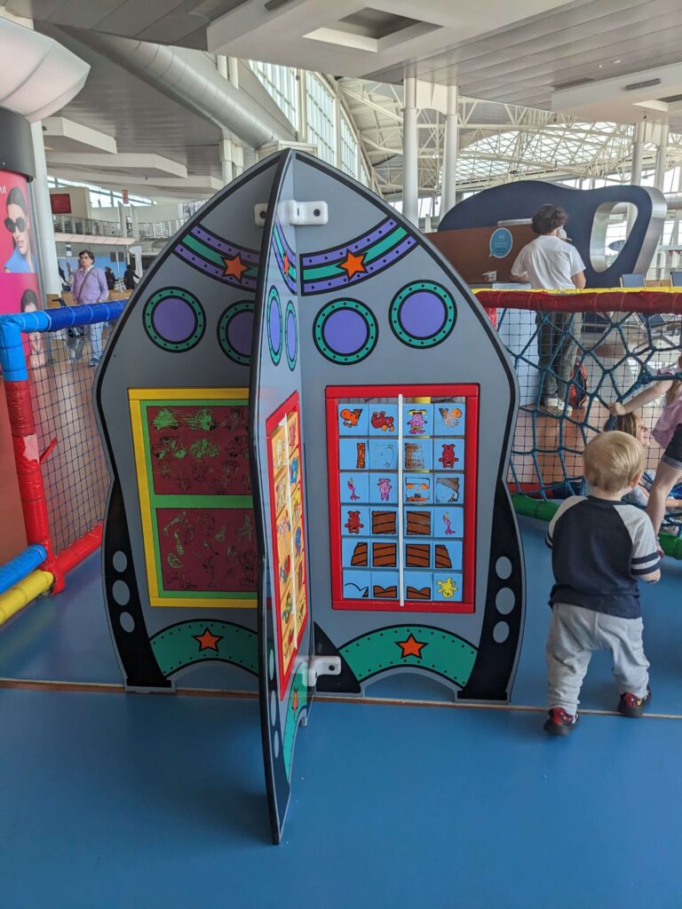 Tout-petit enfant qui s'amuse près d'une structure en forme de fusée colorée dans une aire de jeu de l'aéroport de Porto au Portugal. Les aires de jeux sont l'une des meilleures façon de passer le temps pendant les escales avec des enfants.