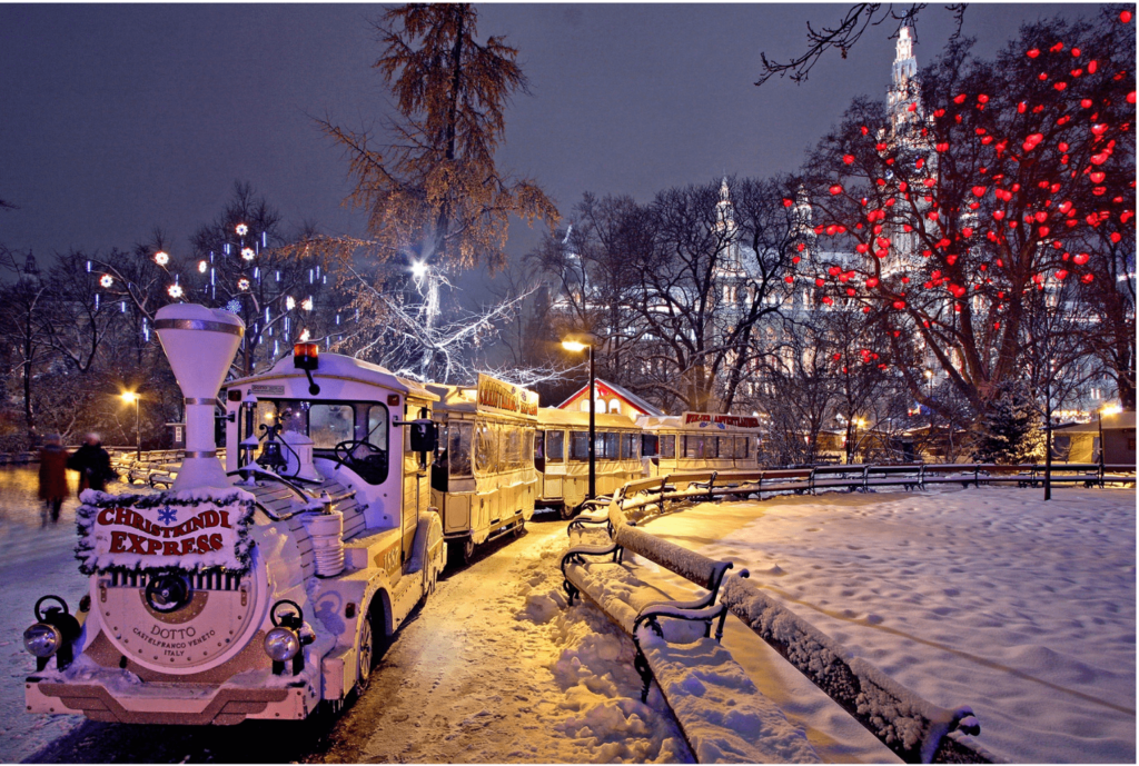 A Christmas Express Train in a snow-covered park at night next to a tree lit up with red Christmas lights at one of Vienna's best Christmas markets for families. 