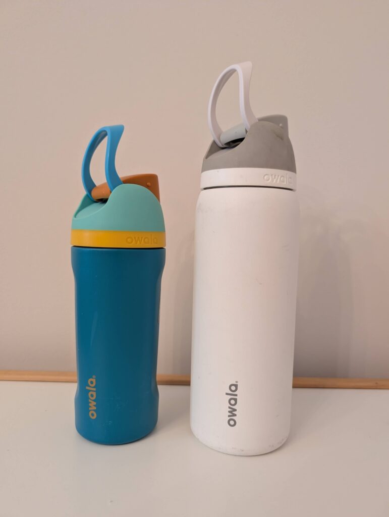 A blue and orange Owala water bottle for kids on the left and a tall white Owala bottle with a gray cap for adults on the right. They keep the water super cold and are worth adding to your hiking essentials for kids checklist!