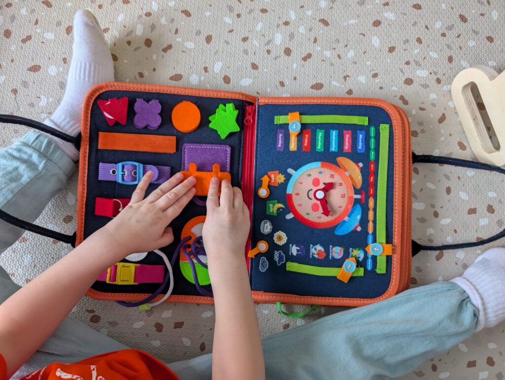 Hands of a toddler playing with a soft busy board on the floor.