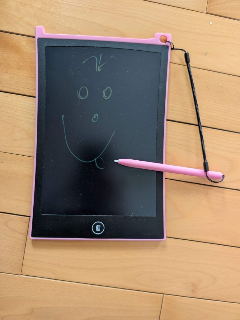 A pink LED doodle board with a smiley face draw on it, laying on a hardwood floor.