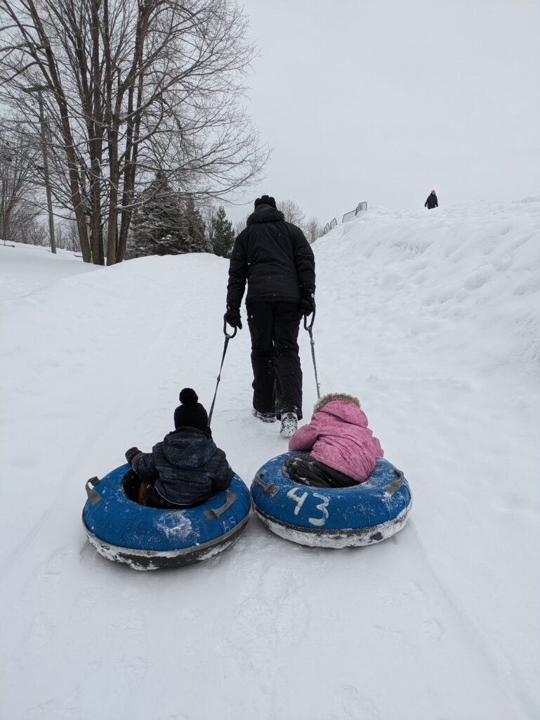 Two kids sitting in a snow tub carried by a man up a snowy hill in Ottawa.