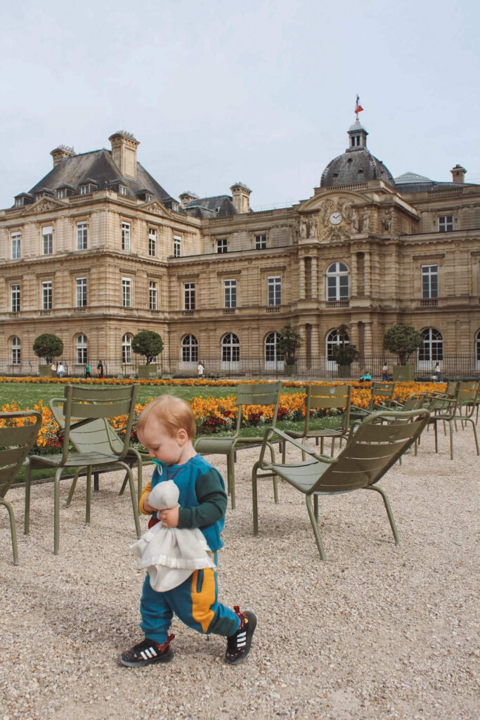 A toddler holding a plush toy walking on a gravel path at the Luxembourg Garden with the Palais du Luxembourg standing in the background. This garden is a fantastic place to visit in Paris both for kids and parents.