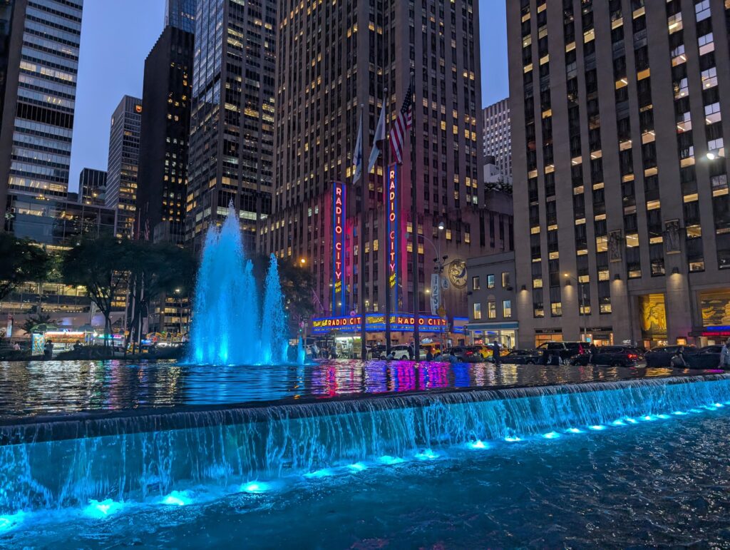 Radio City in New York City at night behind a water fountain lit up in blue.