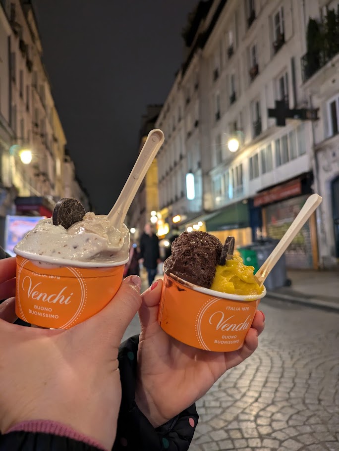 Two hands holding orange cups filled with gelato in Montorgueil in Paris at night.
