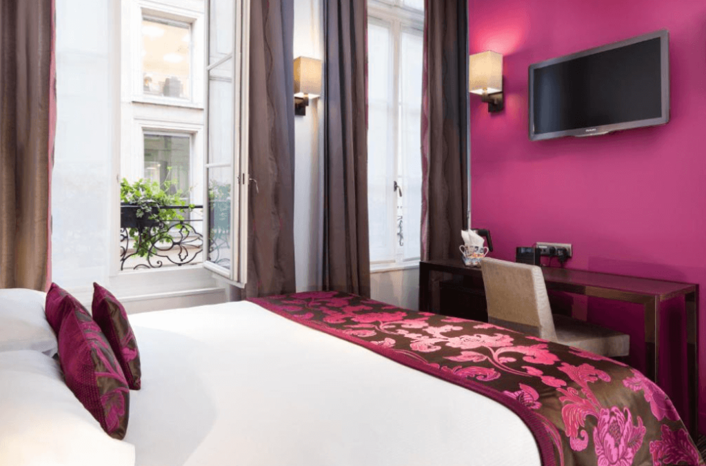 A bedroom at Hotel Paris Louvre Opera, with a double bed against a dark pink wall. Despite its perfect central location, it's one of the best budget hotels in Paris for families. 
