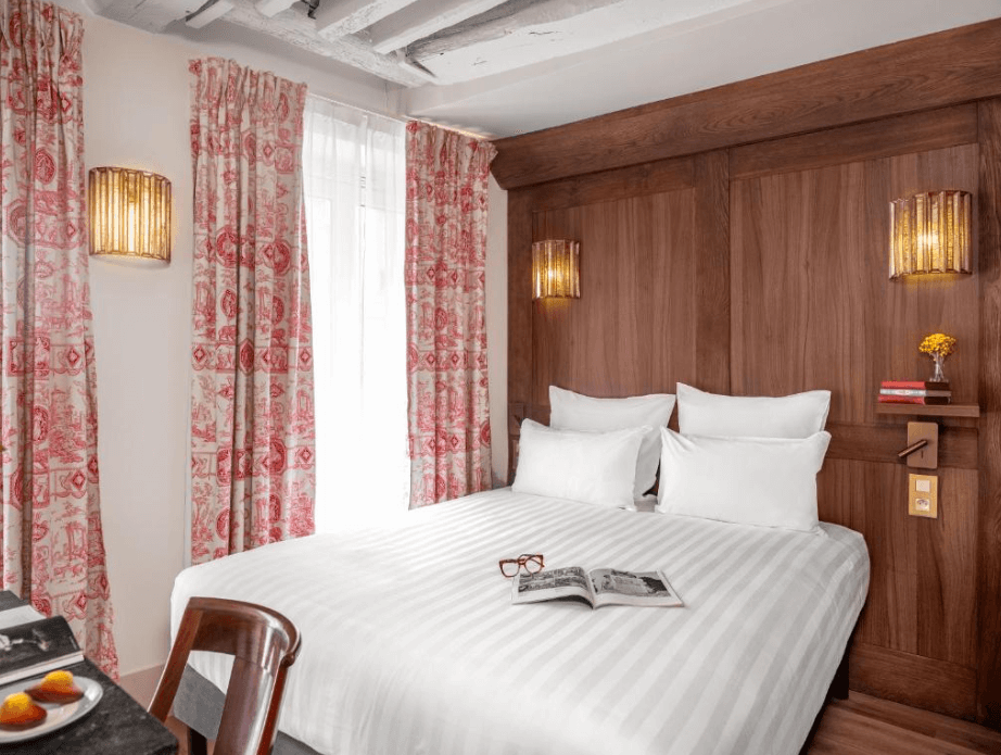 A double bed with white sheets and a white cover next to a red and pink curtains at the Madeleine de Senlis family hotel in Paris. 