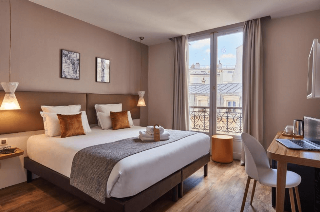 A double bed standing in the  middle of the Magenta 38 by HappyCulture's room, next to a floor-to-ceiling glassed door with views on Paris. The room is spacious and can accommodate families.