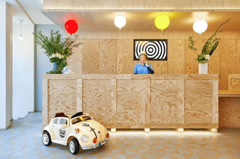 A staff member standing at the wooden lobby's desk of the Hotel Joke from Astotel in Paris. There's a miniature Volkswagen Beetles on the ground where the kids can sit. With lights shaped as balloons, it has a fun family-friendly vibe.