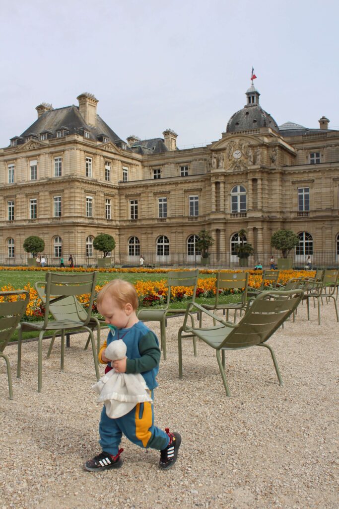 A toddler holding a plush toy is walking in the Luxembourg Garden in Paris on a sunny day. This garden is perfect for kids to run around and for parents to relax while enjoying the views of Paris. 