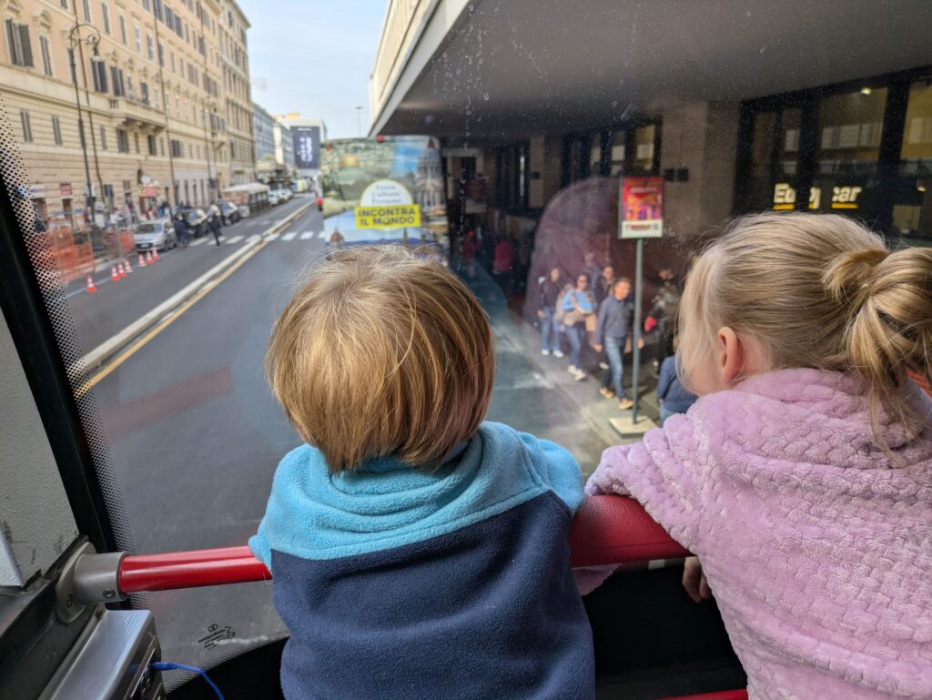 A toddler in a blue vest and a little girl in a purple vest looking at the street through the window from the upper floor of a double-decker bus in Rome, Italy. Hop on hop off bus tours is the best things to rest little legs when visting Rome.