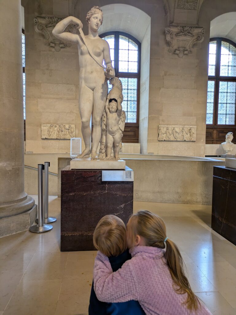 A little girl whispering to her little brother's ear in front of a Greek statue at the Louvre in Paris, France. 