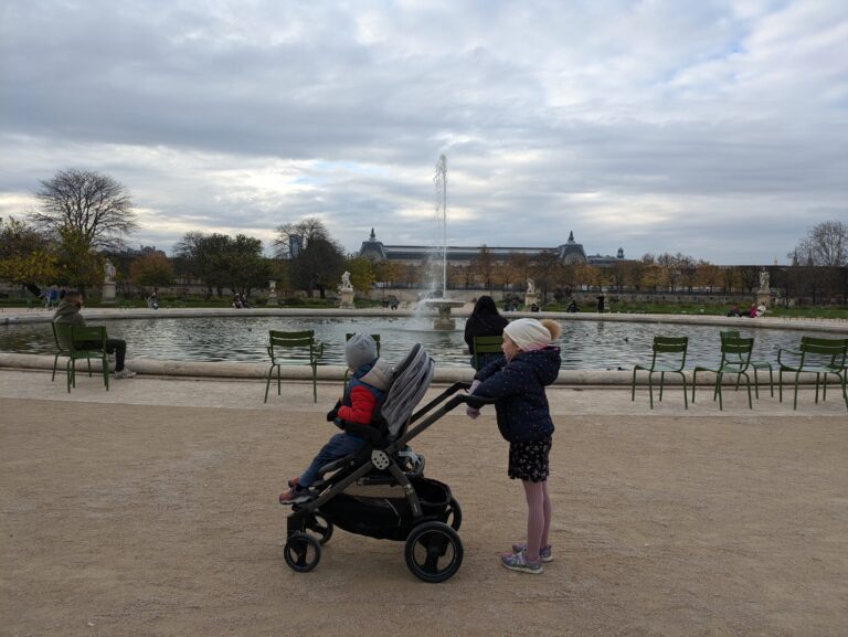 A little girl pushing a stroller with a toddler sitting in it in front of a fountain in the Tuileries Garden in Paris, France, on a cloudy day.