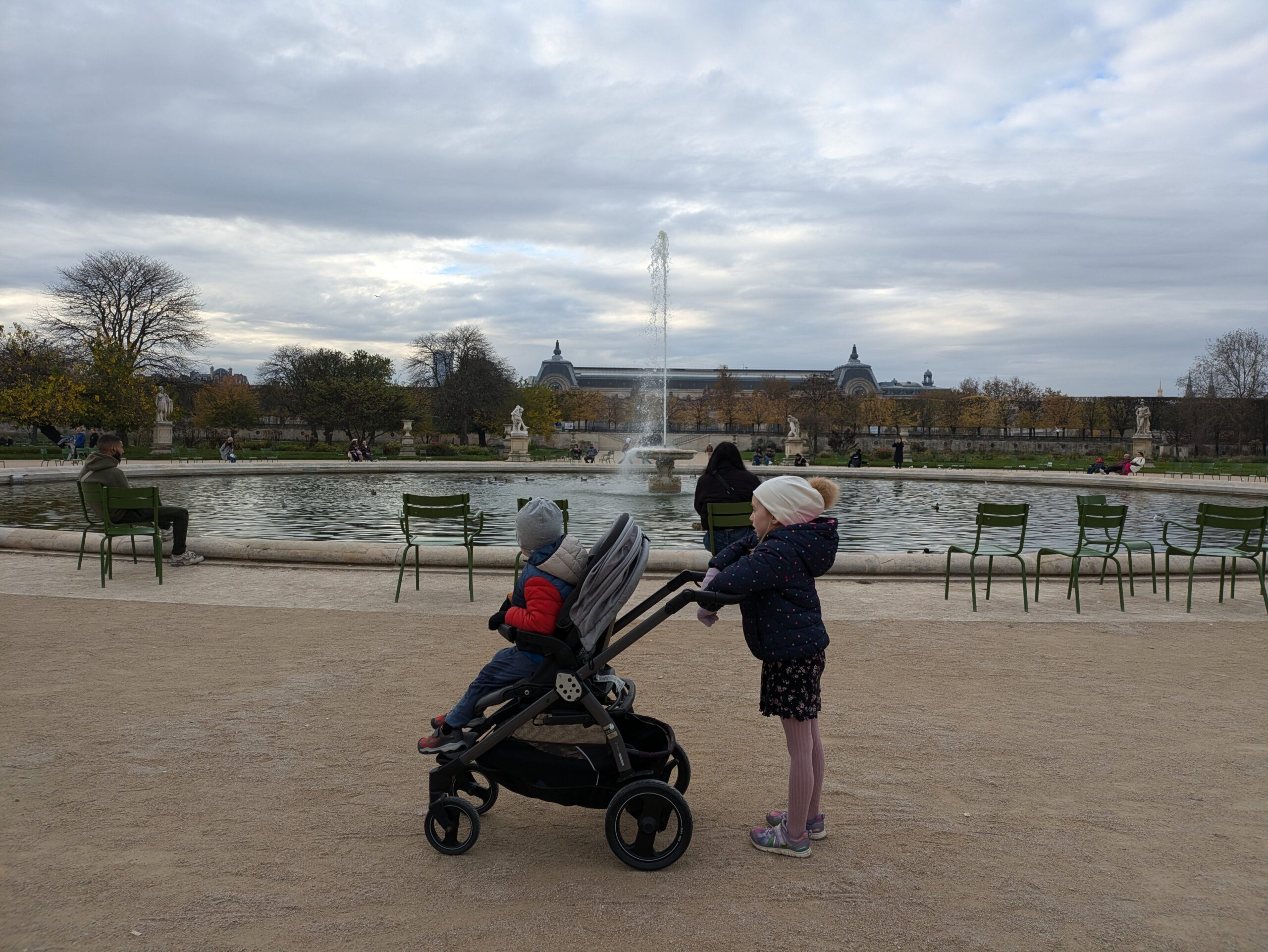 A little girl pushing a stroller with a toddler sitting in it in front of a fountain in the Tuileries Garden in Paris, France, on a cloudy day.