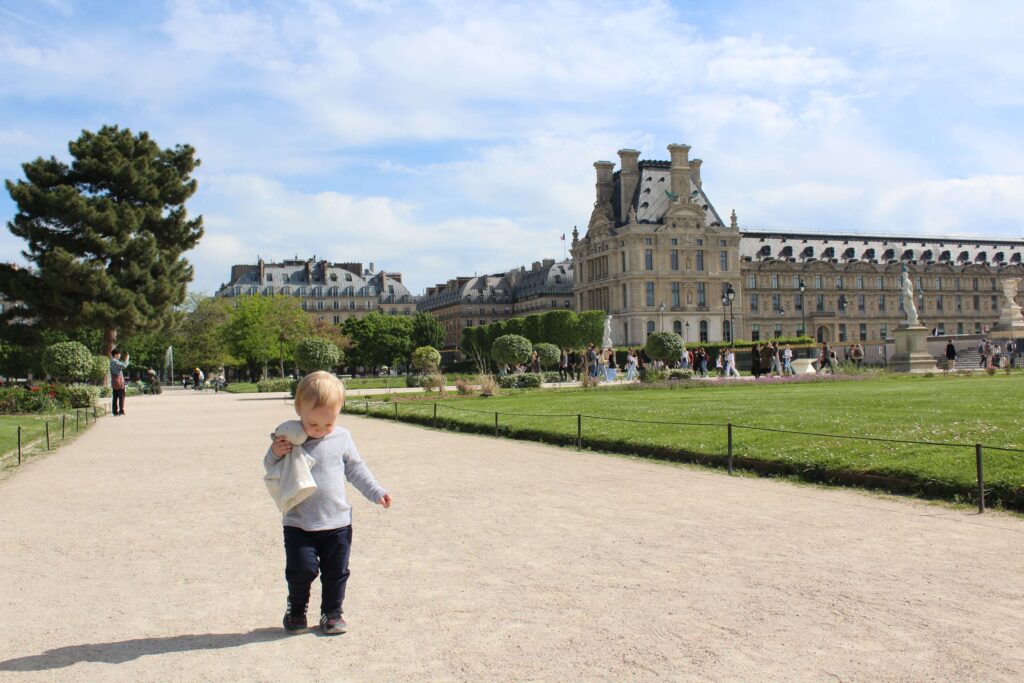 Toddler walking on a gravel path holding a stuffed animal in his right hand in the Tuileries Garden in Paris, France on a sunny day.