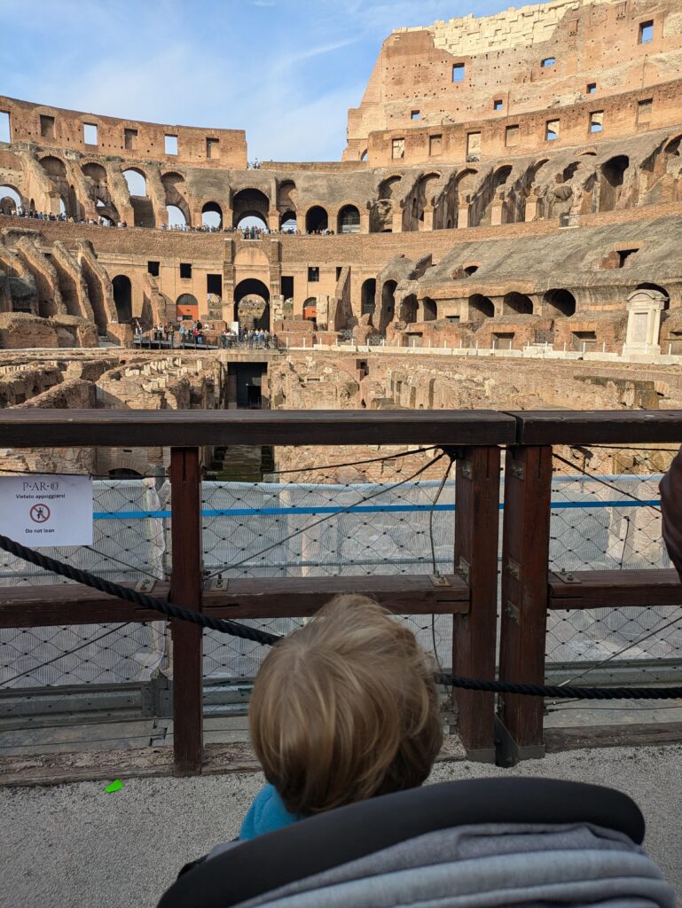 A blond hair toddler sitting in his stroller looking at the Colosseum on the Arena floor on a sunny day in Rome.