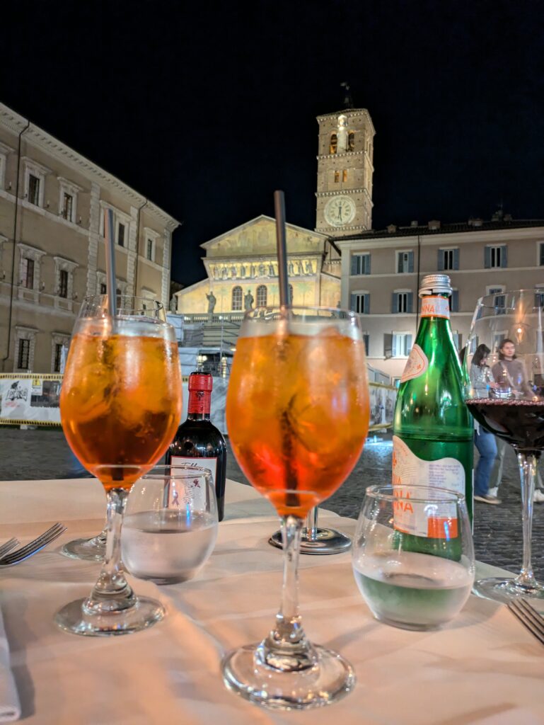 Two glasses of Aperol Spritz on a table of a restaurant in Trastevere in Italy, with Santa Maria Church in the background.