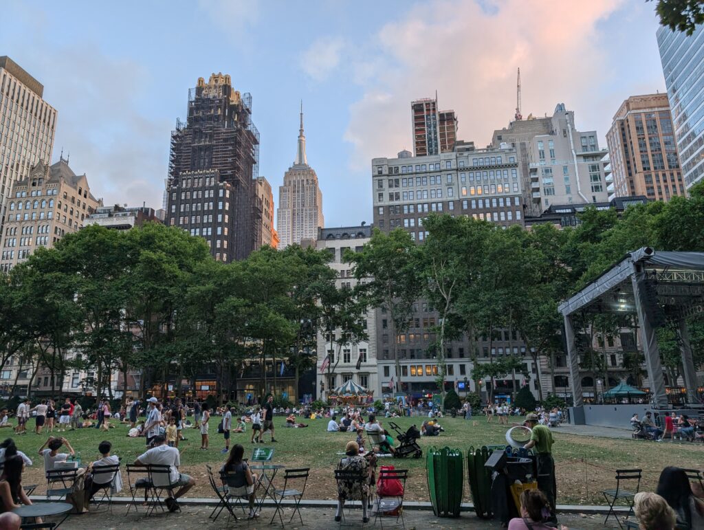 Crowd gathered on the grass at Bryant Park in New York City with the Empire State Building in the background. Not planning breaks in your itinerary is one of the biggest mistakes to avoid in NYC with kids and Bryant Park is one of the best spots to take a break. 