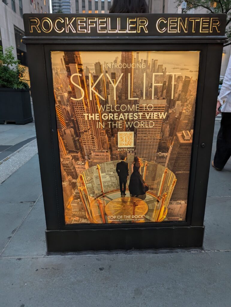 Sign with two people standing at the Top of the Rock observatory deck in NYC.