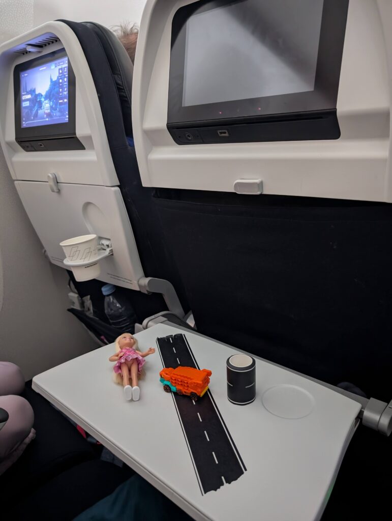 A piece of road tape sticked to the tray with a toy car and a small princess figurine laying on the tray of a kid's seat on a plane. These toys are a must to pack in any toddler's carry-on.