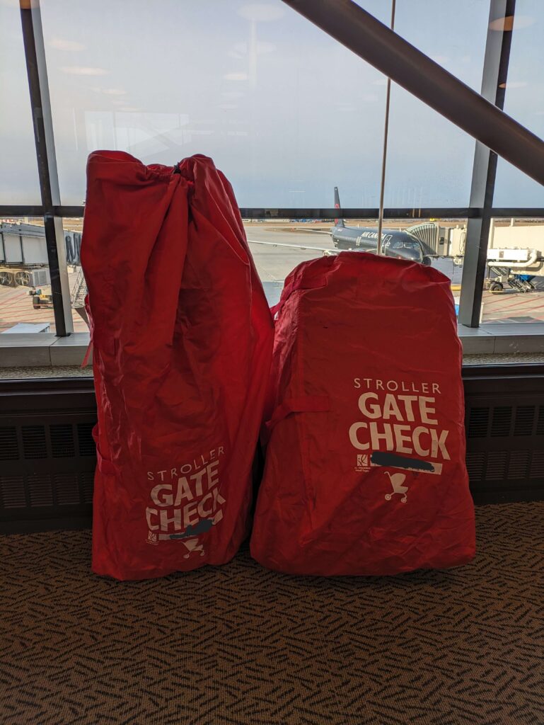 Two red travel stroller bags, from the J.L. Childress Gate Check collection, in front of a window at the airport. This stroller bag is a must for parents wondering how to protect your stroller on a plane. 