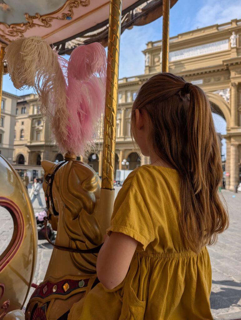 A little girl in a dark yellow dress sitting on a horse of the famous Carousel in Florence. Riding the carousel is hands down one of the coolest and iconic things to do in Florence for families. 