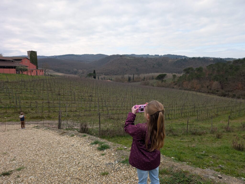 Little girl in a purple jacket and light jeans taking a picture with a kid camera of the Tuscan vineyard near Florence. Wine tours are suprisingly a top family-friendly activity in Florence. 