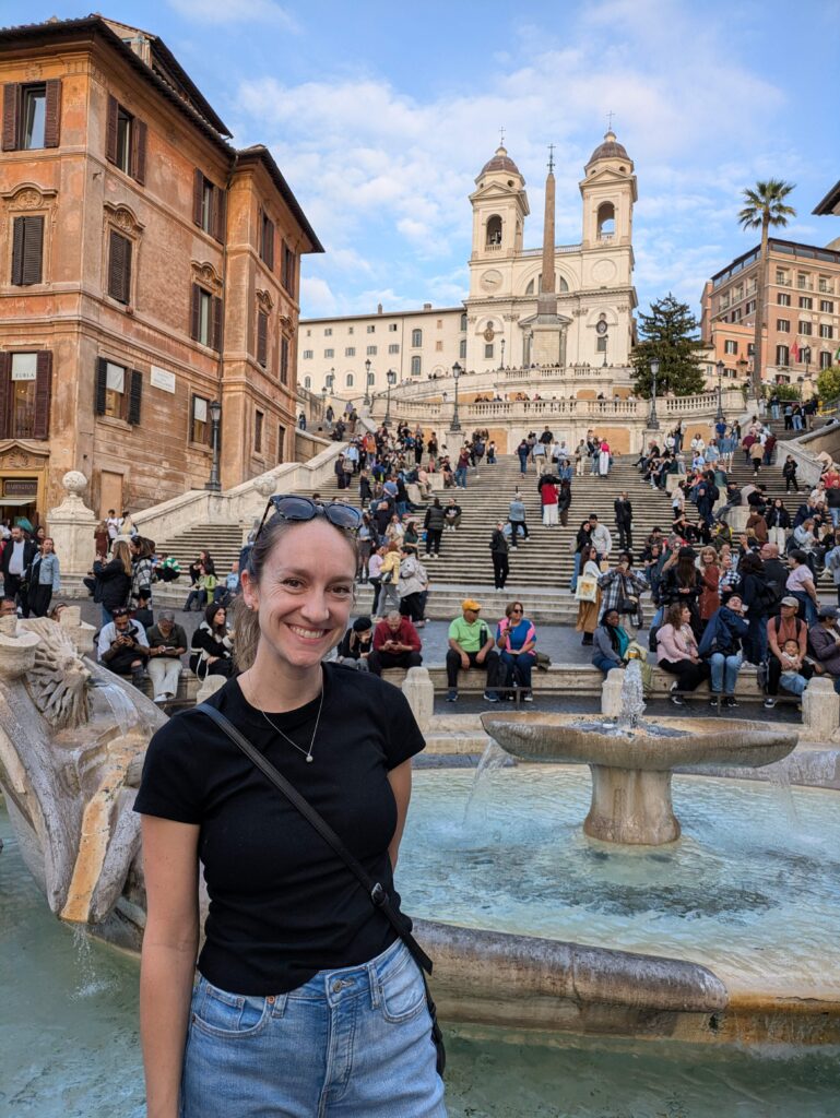 Lady wearing a black tshirt and light jeans smiling and standing in front of the fountain at the bottom of the Spanish Steps in Rome, Italy.