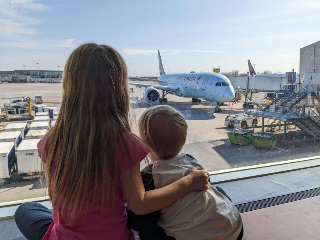 Un tout-petit garçon et sa grande soeur assis sur le bord de la fenêtre à l'aéroport et regardent un avion d'Air Canada pendant une escale. Regarder les avions peut être un bon moyen de passer le temps à l'aéroport quand on voyage en avion avec un tout-petit.