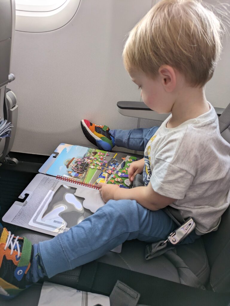 Tout-petit aux cheveux blonds qui s'amuse avec un livre de collants réutilisables Melissa and Doug de la Pat Patrouille sur son siège dans l'avion. Prévoir des jeux calmes est essentiel pour voyager en avion avec un tout-petit.