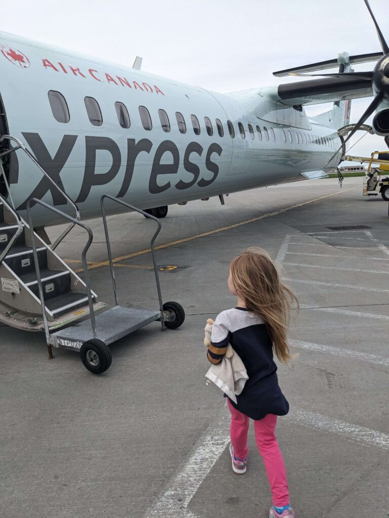 Toute-petite tenant un toutou dans sa main gauche qui s'approche des escaliers pour embarquer à bord d'un avion Air Canada express sur le tarmac.