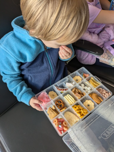 Un tout-petit garçon aux cheveux blonds qui mange une collation dans sa boîte de collations surprise à l'aéroport. Cette boîte de collation est un must à mettre dans le bagage de cabine des tout-petits.