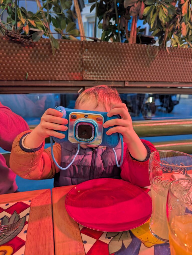 Un tout-petit garçon qui tient un appareil photo Vtech Kidizoom bleu devant son visage. Ce jouet est une excellente façon de divertir les enfants à l'aéroport!